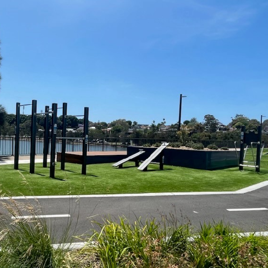 Outdoor fitness equipment on grass in front of a cycleway with water and blue sky in the background 
